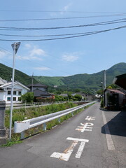 A narrow street in a quiet Japanese village with road markings and power lines under a clear blue sky. Surrounded by green hills and traditional houses, it captures rural charm and peaceful daily life