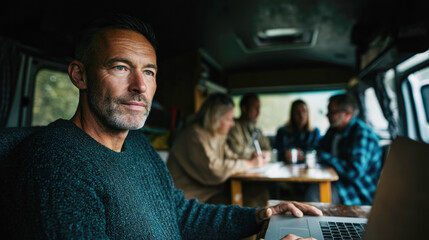 Caucasian adult male in camper working on laptop with friends socializing in background