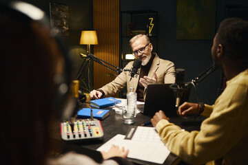 Mature influencer speaking in microphone and discussing book with young people during podcast