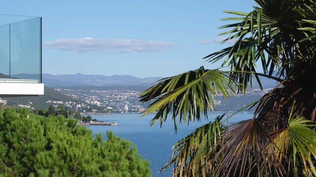 Luxury room with sea view in Lovran, Istria - Croatia. There is a palm tree in the foreground and a mountain range in the background. Seaside balcony on a sunny morning with modern glass railing.