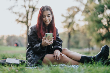 A cheerful young girl with red hair sitting on grass, enjoying her smart phone outdoors.