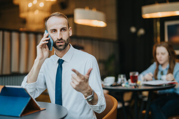Professional man in a formal shirt and tie communicates through a mobile phone inside a stylish cafe environment during the work day. Background blurred with a sense of activity.