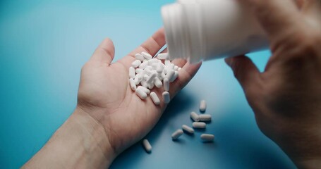 Close-up slow motion shot of white pills being poured into a hand against a solid blue background. Medical and pharmaceutical concept with clean composition. - Powered by Adobe