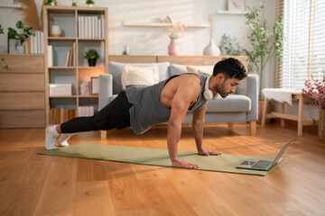 Muscular man wearing headphones in high plank pose on mat, engaging in strength training while watching an online video at home.