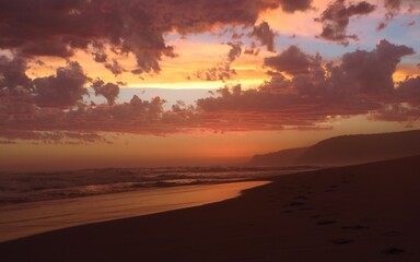 Obraz premium A tranquil beach scene at sunset with vibrant clouds reflecting warm hues, serene waters, and a peaceful atmosphere in Nature's valley, South Africa