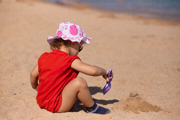Cute baby girl in fun hat and sun glasses playing on the beach in sand with toy paddle. Builds a sand castle on sea background. Summer sunny vacations