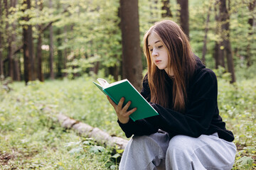 Teenage girl reading a book while sitting on a log in the forest, copy space.