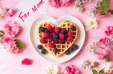 Heart-shaped waffle with berries and jam on white plate with Mother's Day cards and flowers








