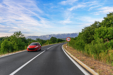 A red car drives along a winding mountain road surrounded by lush greenery, under a bright blue sky with a visible no overtaking traffic sign.