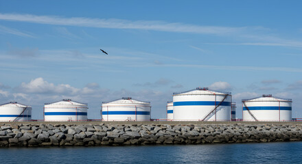 Fototapeta premium Row of White and Blue Industrial Storage Tanks at a Coastal Oil Refinery Plant Under a Clear Sky Seascape