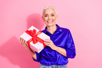 Smiling senior woman holding a gift box with a vibrant red ribbon against a bright pink background