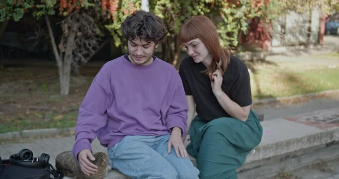 A young couple sits on a concrete ledge, petting a stray cat and talking. The scene evokes feelings of kindness, care, and connection.
