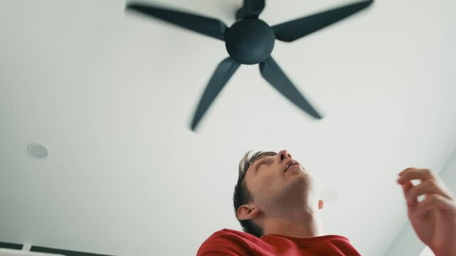 Exhausted man sitting under ceiling fan at home, cooling off during hot summer weather, suffering from heat, high temperature. Young male turn on electric fan to cool off under ventilator