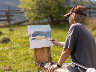 Female artist creates landscape painting in a tranquil meadow surrounded by majestic mountains...