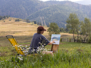 Female artist creating a landscape painting in a picturesque meadow surrounded by mountains during a sunny day