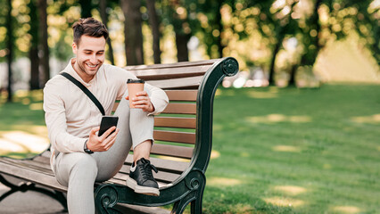 Fototapeta premium A young man relaxes on a bench in a vibrant urban park, sipping coffee from a cup and looking at his smartphone. The sun shines through the trees, creating a lively atmosphere, copy space