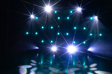 A vibrant stage setup features intense white spotlights and teal-blue LED lights arranged in a...