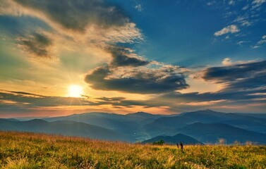 Golden sun rays pierce through dramatic clouds over layers of misty mountains, casting a warm glow across the grassy hilltop at sunset.