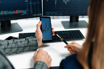 Cropped pointing hand with pen in businessman, checking with holding smartphone by woman investor online website program application with dynamic stock market at home office at back side. Infobahn.