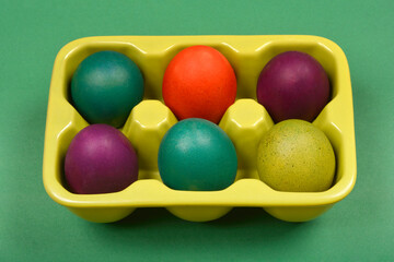 A slightly angled top-down shot shows a light green ceramic egg carton with six colorful Easter eggs on a solid green background