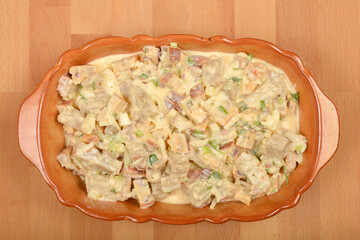 A top-down view shows an oval, light brown ceramic baking dish filled with a creamy mixture of cubed bread, vegetables, and cheese, prepared for a strata