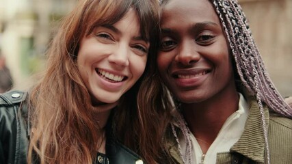 Two young multiethnic women are embracing and smiling joyfully ouddoors, both looking at the camera. Lesbian couple spending time together