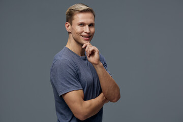 Confident young man with short blonde hair wearing a casual blue t-shirt smiling and touching his chin, posing thoughtfully against a plain gray background. Studio portrait representing positivity and