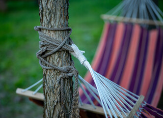 Close-up of hammock ropes tied around tree trunk with striped fabric in the background. Outdoor...