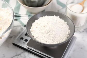 Digital kitchen scale with bowl of flour and other products on white marble table, closeup