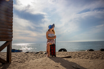 young girl, sitting on a suitcase, on the beach by the sea, looking away, long white hair, blue hat, sundress, bare legs, 19 years old, sky with clouds, calm, vacation, travel, relaxation