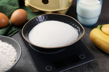Digital kitchen scale with bowl of sugar and other products on grey table, closeup