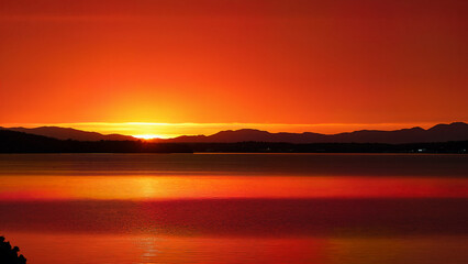 Obraz premium Sunset with vibrant orange and red hues reflecting on lake, mountains silhouetted in the background. Perfect for nature, relaxation, and landscape inspiration searches.