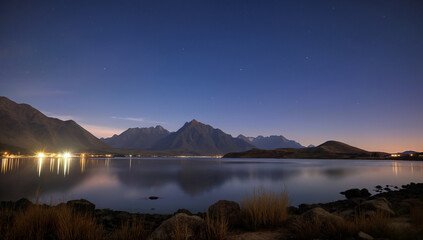 Fototapeta premium Landscape with lake and mountains at night. City and stars reflecting in water. For illustrating nature, astronomy, travel scenery concepts.