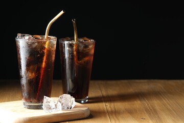 Refreshing cola with ice cubes and straws in glasses on wooden table against black background,...