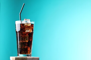 Refreshing cola with ice cubes and straw in glass on stand against turquoise gradient background,...