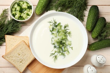 Tasty cucumber soup in bowl and ingredients on light wooden table, flat lay