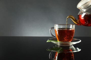 Pouring freshly brewed tea from teapot into cup at dark mirror table, closeup. Space for text