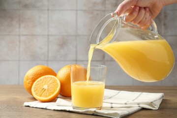 Woman pouring fresh orange juice from jug into glass at wooden table against grey tiled wall, closeup