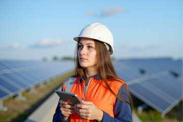 Young Female Technician in Hard Hat with Tablet Observing Outdoor Solar Farm, Profile View.