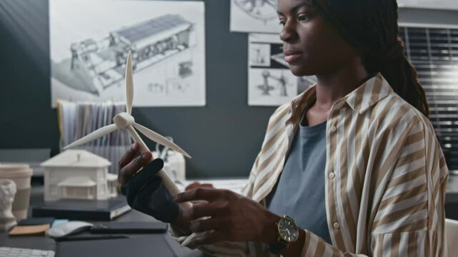 Side shot of young female Black innovator observing small wind turbine model and drawing digital solar power system on tablet at engineering workspace in sustainable technology office