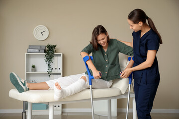 Female doctor helping woman with broken leg and crutches to stand up from couch in clinic