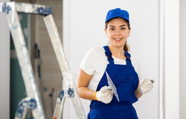 Portrait of smiling young female plasterer wearing blue overalls and cap standing with finishing...