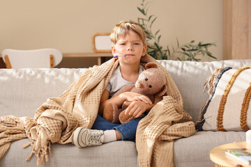 Little boy ill with chickenpox and toy bear measuring temperature on sofa at home