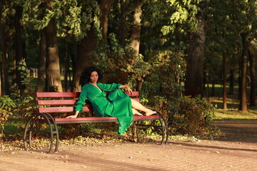 Slender barefoot curly woman in green dress sitting on a bench in park full length