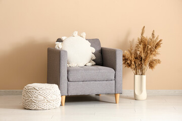 Armchair with pillow, pampas grass in vase and pouf near beige wall in room
