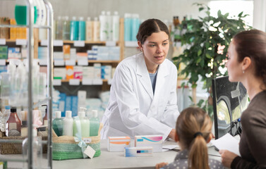 Obraz premium Professional young pharmacist using computer to check medication stock, politely explaining treatment options, and processing order for female customer with tween daughter
