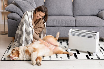 Young woman with blanket and Australian Shepherd dog sitting on floor near heater at home
