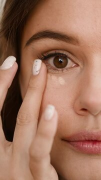 Focused lady blends liquid concealer under eyes with finger to cover dark circles. Young woman prepares facial skin for professional makeup closeup