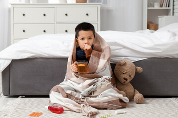 Ill little Asian boy with plaid and cup of tea coughing in bedroom