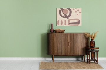 Chest of drawers, pampas grass in vase on stool and picture on green wall in room